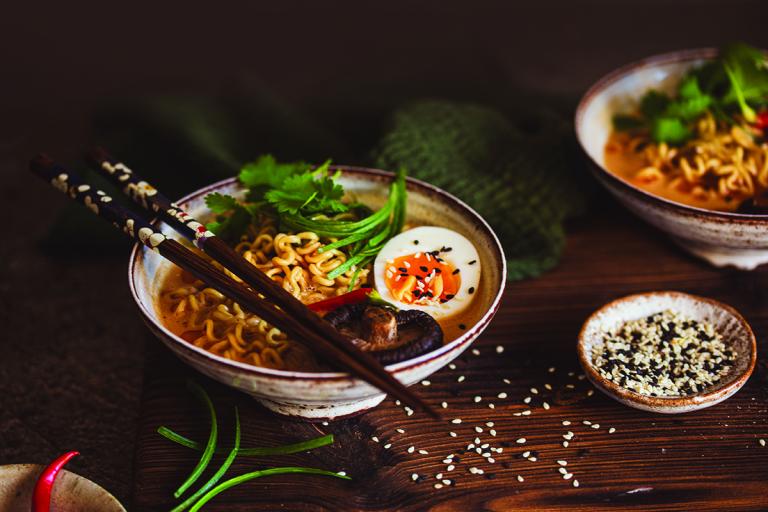 A bowl of ramen noodles with half a hard-boiled egg and cilantro in a small white bowl with brown chopsticks resting on top.